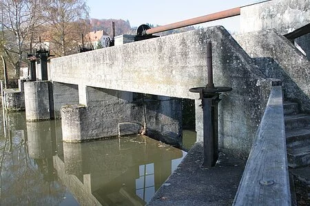 Das massive Betonwehr steht im Mittelpunkt. Rechts sind Treppenstufen hinter einer Mauer zu erkennen. Dahinter kann man vereinzelt ein paar Häuser erkennen.