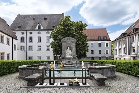 Zentrum des Bildes ist eine Ädikula mit einer Steinstatue, die die heilige Maria, die Patrona Bavaria, mit Kind darstellt. Unmittelbar davor befindet sich das rechteckige Brunnenbecken, welches von einer kniehohen Stein-Mauer begrenzt wird.
Dahinter stehen ein großer Baum und niedrige Hecken, die den Kiesplatz, auf dem der Brunnen steht, einsäumen.
Im Hintergrund befinden sich verschiedene mehrstöckige Häuser in hellen Farben.
