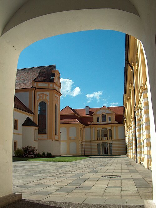 Ein gemauerter Torbogen umrandet das Bild, durch den man in den Innenhof gelangt. Der Hof zeichnet sich durch Grünflächen und den hellgepflasterten Boden aus. Um ihn herum stehen mehrere Gebäude. Sie sind in weiß und gelb gehalten. Auf der linken Seite erstreckt sich ein Teil des Kirchenschiffs. Auf der rechten Seite und im Hintergrund sind die Gebäude, die Räume der Maria-Ward Realschule beherbergen.