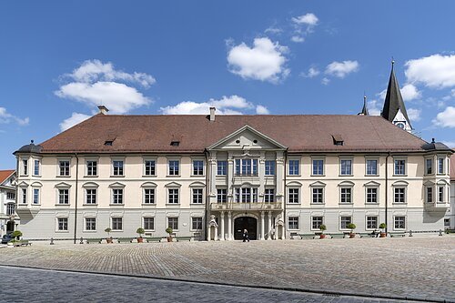 Über das ganze Bild erstreckt sich die Vorderseite der dreistöckigen Fürstbischöflichen Residenz. Der mittlere etwas vorgezogene Gebäudeteil hat ein großes zweiflügeliges Tor, das links und rechts von zwei Wachhäuschen aus Stein flankiert wird. Unmittelbar darüber befindet sich ein Balkon.
Die Fassade der oberen beiden Stockwerke ist cremefarben das Erdgeschoss ist hellgrau gestrichen. Das Dach ist mit dunkelroten Ziegeln eingedeckt.