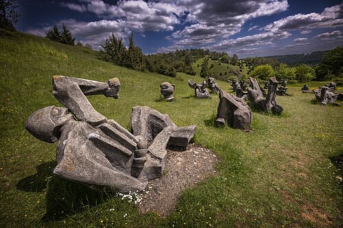Die Fotographie zeigt ein Skulpturenfeld, welches auf einer Wiese in Hanglage gelegen ist. Die abstrahierten, menschenartigen Figuren sind aus Beton gegossen und stellen Kämpfer und Opfer in verschiedenen Positionen dar. Die Gesamtheit des Figurenfeldes erinnert an ein Schlachtfeld. Der Hintergrund zeigt eine karge Berglandschaft.