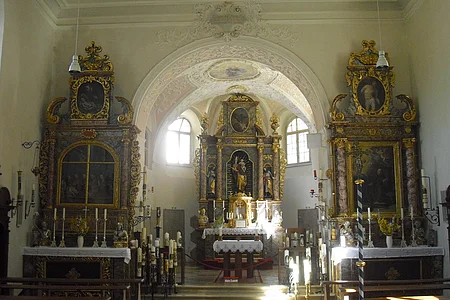 Innenraum einer Kirche mit barocken Altären, verziert mit Gemälden und goldenen Ornamenten. Sonnenlicht fällt durch ein Fenster.