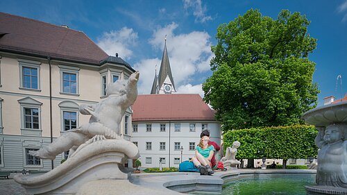 Wanderer am Residenzplatz in Eichstätt Paar sitzt am Rand eines Brunnens mit Steinfiguren vor historischen Gebäuden und einer Kirche bei blauem Himmel.