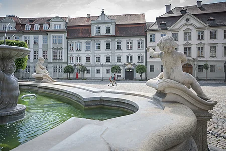 Brunnen mit Puttenfiguren im Vordergrund und historische Gebäude mit roten Dächern im Hintergrund, zwei Personen gehen.