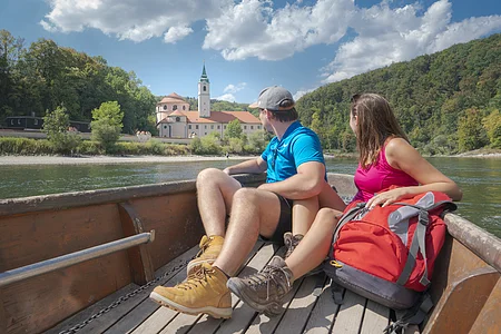 Paar sitzt in einem Boot auf einem Fluss und blickt auf ein Gebäude mit Turm und Wald im Hintergrund.