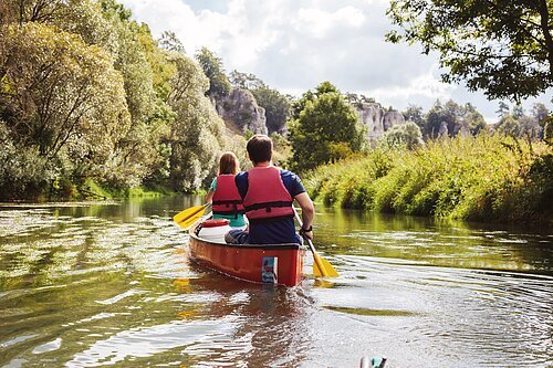 Bootwanderer auf der Altmühl Zwei Personen mit roten Schwimmwesten paddeln in einem roten Kanu auf einem Fluss, umgeben von grüner Natur.