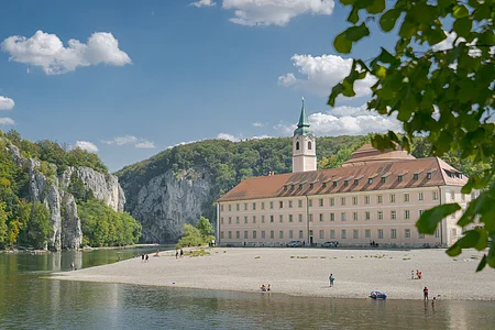 Flussufer mit Kiesstrand, altes Gebäude mit Turm und bewaldete Felsen unter blauem Himmel mit Wolken