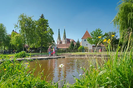 Park mit Teich, Wiese, Bäumen und Menschen vor historischen Gebäuden mit zwei Türmen bei blauem Himmel