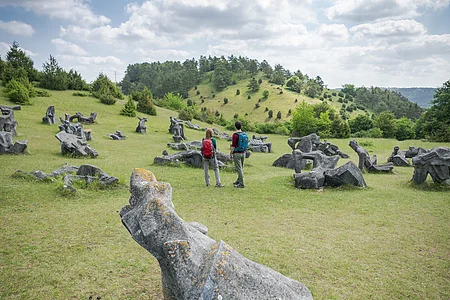 Zwei Personen mit Rucksäcken stehen auf einer Wiese mit verstreuten großen Steinfiguren unter bewölktem Himmel.