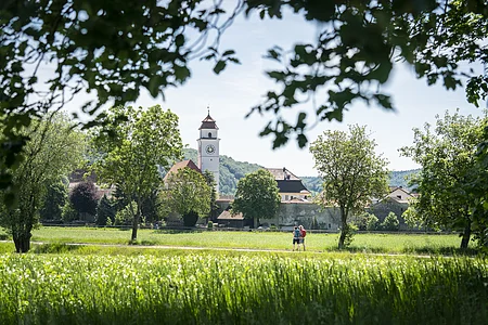 Zwei Personen gehen auf einem Weg durch eine grüne Wiese, im Hintergrund ein Kirchturm und Bäume.