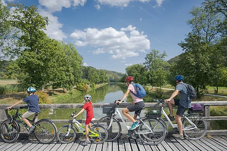 Vier Personen mit Fahrrädern auf einer Holzbrücke mit Blick auf einen Fluss und grüne Landschaft.