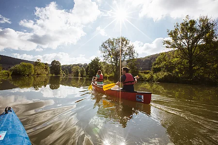 Zwei Personen paddeln in einem roten Kanu auf einem ruhigen Fluss bei sonnigem Himmel mit Wolken.