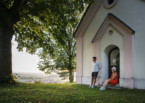 Wanderer auf der Monheimer Alb Mann lehnt an Hauswand, Frau sitzt in Türbogen, große Bäume, Dorf im Hintergrund bei Tageslicht.