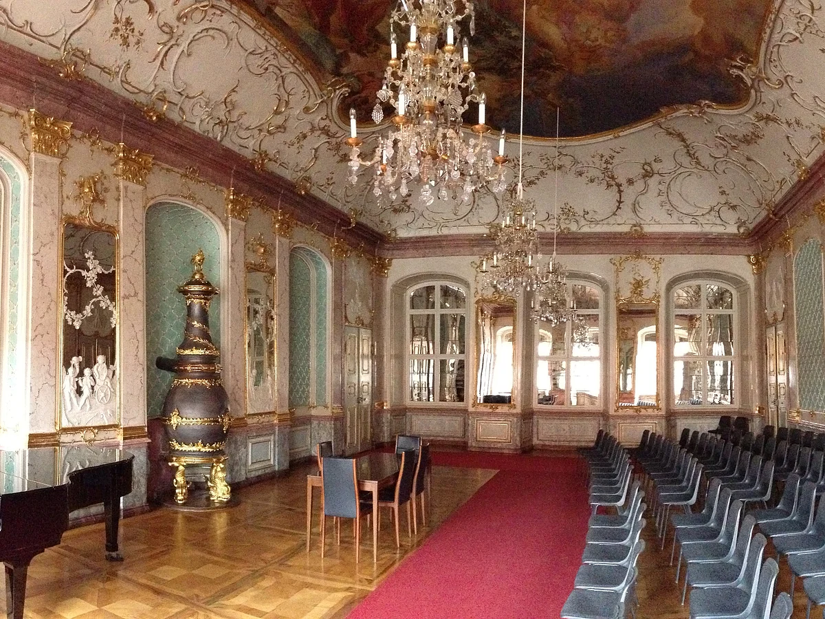 Prunkvoller Saal mit Kronleuchtern, roten Teppich, Klavier, Stühlen und goldverzierten Wänden und Decke.