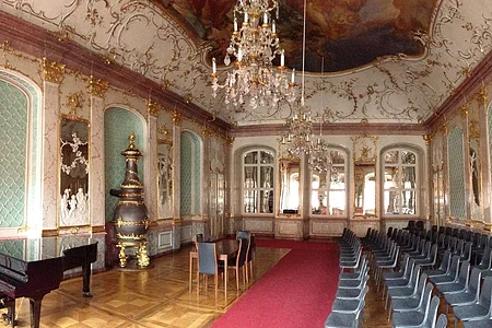 Prunkvoller Saal mit Kronleuchtern, roten Teppich, Klavier, Stühlen und goldverzierten Wänden und Decke.
