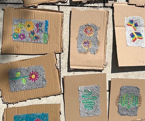 Mehrere Hand geschöpfte Papierstücke mit Blumen- und Schmetterlingsmotiven liegen auf braunen Kartonstücken auf einem Steinboden.