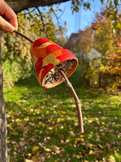Hand hält bemalten Blumentopf mit Vogelfutter an einem Ast vor unscharfem Gartenhintergrund im Herbst
