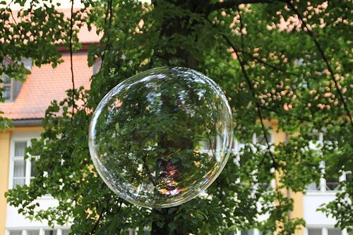 Eine riesige Seifenblase schwebt in der Höhe, hinter ihr die grünen Äste eines Baumes. Durch diese ist ein Gebäude zu erkennen.