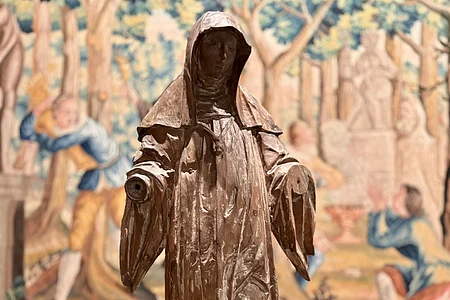 Vor einem Wandteppich mit vielen Baumverzierungen steht eine hölzerne Statue.