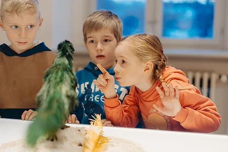 Drei Kinder beobachten aufmerksam ein grünes, federartiges Objekt hinter einer Glasvitrine.