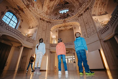 Vier Kinder stehen in einem historischen Raum und blicken zur verzierten Decke mit Fenstern und Malereien.