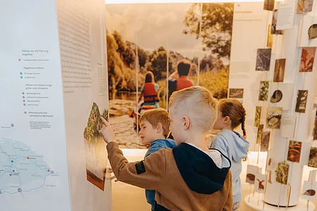 Drei Kinder betrachten eine Ausstellungstafel in einem Museum mit Natur- und Landschaftsbildern.