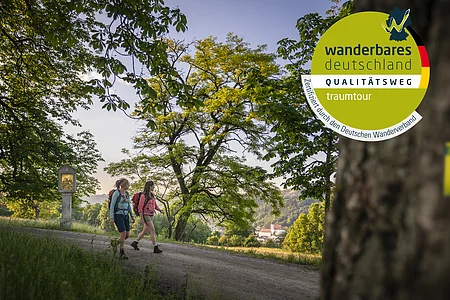 Zwei Wanderinnen auf einem Weg unter Bäumen, Wanderweg-Markierung an Stamm im Vordergrund