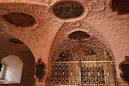 Decke mit Stuckverzierungen und bemalten Medaillons über einem goldverzierten Gitter in einem historischen Innenraum.