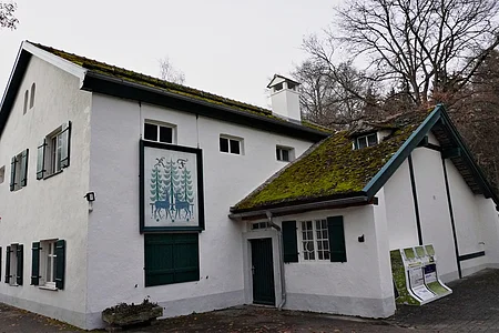 Weißes zweistöckiges Haus mit grünem Fensterladen und Moos auf dem Dach, im Herbstwald gelegen.