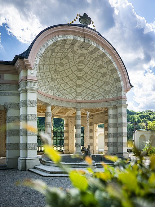 Auf dem Bild ist der mittlere und größte Pavillon des Hofgartens. Der Pavillon ist rund und nach vorne hin geöffnet. Ein Brunnen befindet sich im Zentrum. Dahinter stehen mehrere weiß, grau gemusterte Säulen. Die Kuppel ist innen mit einem mosaikartigen Muster aus grauen und weißen Elementen geschmückt. Das Dach wird von einer vasenähnlichen Figur geziert.
Bildbeschreibung ehemalige Kapuzinerkirche (innen)