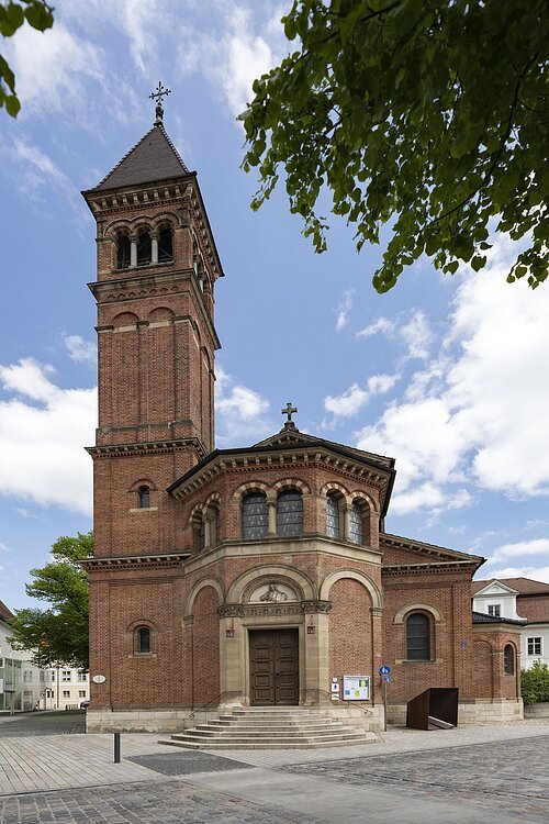 Das Bild zeigt eine Kirche aus roten Ziegeln. In der Mitte führt eine helle Steintreppe zum Kircheneingang. Über dem Portal befindet sich ein Abbild von Jesus. Auf der linken Seite der Kirche erhebt sich ein Glockenturm mit schwarzem Dach, der fast doppelt so hoch wie die Kirche ist. Darauf steht ein Kreuz.